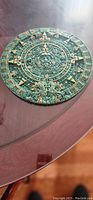 Top view of a round green stone Mexican calendar with intricate carved Aztec or Mayan designs and gold-colored details, diameter approximately 30 cm.