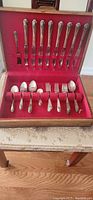 Wooden box set of silverware with red velvet lining. Contains 8 knives placed vertically at back, 4 forks, 2 soup spoons, 4 smaller spoons at the front row with several empty slots.
