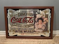 Frontal and angled views of the vintage Coca-Cola advertising mirror showing detailed artwork and wooden frame.