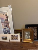 Six framed artworks and matted photographs arranged on wooden table and floor, showing variety in size and style