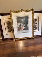 Three framed artworks leaning against a wall on hardwood floor. Largest artwork with ornate gold frame in center and two smaller dark framed artworks behind.