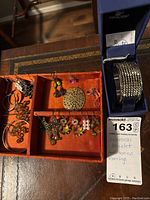 Photo of a Swarovski bracelet in original box beside an orange organizer tray with multiple pairs of various style earrings including hoops, drop bead, filigree medallion, butterfly and pearl earrings.