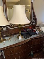 Two brass lamps shown on wooden dresser with beige fabric lampshades, also glasses and decor visible