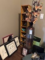 Photo showing wooden shelving unit with various small decor pieces, tall vase with artificial orchid flowers, and multiple framed prints.