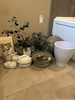 Wide view showing various ceramic decor items including dark green ceramic set and white bathroom ceramics grouped on floor against a beige wall.