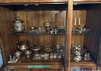 Full display of various EP and stainless steel serveware items arranged on wooden shelf inside glass cabinet.