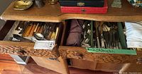 Open wooden drawers filled with varied silverware cutlery pieces with some visible knives, forks, and spoons, along with handle diversity including wood and metal handles.