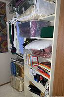 Wide view showing stacked bedding sets, pillows, plastic containers with fabric or similar items, books on white shelving, and small electronic devices.