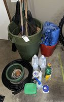 Full view of assorted gardening items including tools, pots, coiled hose, chemicals, and accessories arranged on floor near large green trash bin