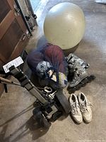Golf push cart, exercise ball, mesh bag with balls, inline skates, and white athletic shoes seen side by side on concrete floor.