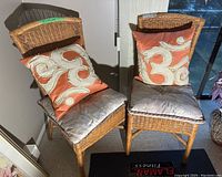 Two wicker chairs each with silver-gray seat cushions and orange decorative pillows with cream scroll patterns placed indoors.