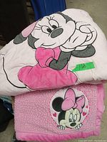 Photo showing two Minnie Mouse crib blankets stacked, one with a large Minnie Mouse lying down image, the other textured pink with a circular Minnie Mouse face graphic.