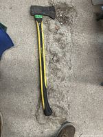 Full view of the used axe laying on a concrete surface showing the yellow and black handle and metal head with a green sticker marked '182'.