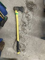 Photo of a used axe with a yellow and black handle resting on a concrete surface