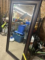 Full view of the large rectangular mirror leaning against a wall in a cluttered storage area, showing the reflective surface and dark frame.