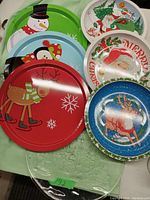 Image showing six round Christmas-themed serving trays arranged on a green tablecloth. Designs include snowman, penguin, Santa Claus, reindeer, and Christmas tree motifs.