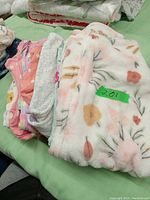 Stack of used baby clothes laid on a green bedspread showing various pastel-colored soft garments including fleece and cotton pieces with floral and animal print patterns.