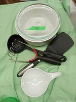 Photo showing two black-handled kitchen utensils (slotted spoon and potato masher), one black-handled spatula, a white ceramic small bowl with a pouring handle, and a round white plastic container with a clear lid.