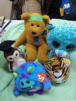 Photo showing 5 stuffed animals. Central is a brown teddy bear with a blue ribbon. Surrounding it are four Ty Beanie Boos style plush toys including a blue and green spotted large-eyed plush, a brown and white penguin, a blue and purple monster, and a tiger-striped tiger plush with large eyes and Ty tags.