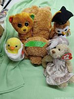 Photo showing five stuffed animals arranged on green fabric including a large brown bear, yellow chick in egg, teddy bear in dress, orange-brown dog, and brown owl with hat.