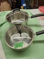 Two stainless steel pots with glass lids on a green surface. Both have black handles and glass lids with black knobs. Signs of light use visible inside pots.