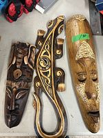Front view of three wooden wall hangings showing two tribal masks and one carved lizard design