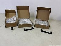 Three cardboard boxes with new cabinet hardware inside plastic bags; two types of hardware visible—a black thick rectangular handle and a small round knob.