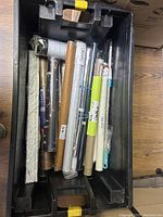 Black plastic tote containing multiple rolled items including posters, adhesive films, tinting films, and poster holders; tote shows signs of wear