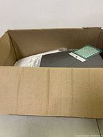 Open cardboard box showing multiple items inside, including a dark gray square item wrapped in plastic and other packaged items.