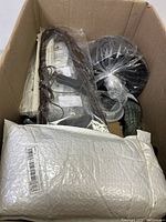 Overview of large cardboard box filled with assorted items including black coiled hose, brown braided cord, green textured sprayer, and multiple boxed goods