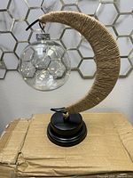 Clear glass orb with LED string lights hanging from crescent moon-shaped twine wrapped stand on a black circular base, shown unlit