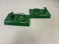 Two green painted metal tractor bucket hooks attached to rectangular metal plates, placed side by side on white surface.