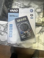 Photo of boxed VARO digital multimeter showing front with model image, product use icons, and 3 year guarantee badge.