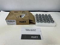 Set of 20 silver chrome wheel lug nuts arranged on foam padding with a damaged cardboard box and a user manual placed in front