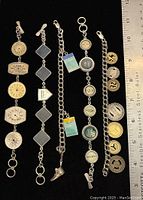 Photo showing five charm bracelets laid out flat beside a ruler for size reference. Bracelets have various charms including circular, diamond-shaped, and Monopoly game related themed charms.