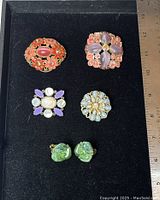 Front view of four decorative brooches and one pair of green art glass clip-on earrings on black background with ruler for scale.