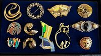 Top view of 13 assorted modern brooches arranged on a dark blue velvet background, varied styles and colors, mostly gold and silver-tone.