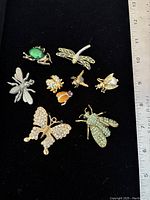 Photo of front side of the 9 insect-shaped brooches and pendants on black background with ruler for scale.