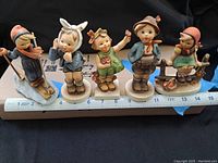 Front view of five Hummel porcelain figurines placed in a row on a measuring ruler, showing detailed front of each child figure in varied poses and clothing