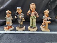 Front view of four Hummel figurines on black backdrop with ruler for scale.