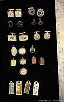 Photo of many sterling silver cuff links and pendants arrayed in black display tray with ruler for scale, showing various themed designs including crossword puzzles, rotary dial, yardstick, typewriter keys, and stadium seats.