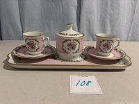 Front view of two tea cups on saucers flanking a covered sugar bowl, all set on a rectangular tray with pink and white floral design and gold accents.