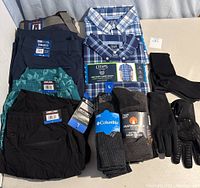 Men's clothing lot of multiple items including dress shirts, casual shirt, pants, socks, and gloves laid out on a surface
