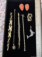 Top-down view of jewelry lot including bracelets, necklace with pendant, coral rose earrings, and pins arranged on black felt with ruler for scale.