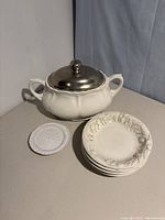 Soup tureen with pewter lid next to 2 small embossed white plates and a round embossed ceramic coaster.