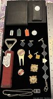 Full lot image showing wallet, bottle opener with red handle, various cufflinks, keychains, character pins, and bracelets arranged on black velvet surface alongside measuring ruler.