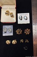 Top-down view of assorted Trifari earrings including boxed gold-tone ribbed clips, oval white center studs, floral silver-tone studs, gold-tone cluster designs, and smaller clip-on earrings alongside a ruler for scale.