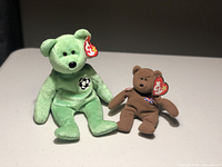 Two Ty Beanie Baby bears on a light-colored table: green sitting soccer bear with black embroidered soccer ball detail and brown Britannia bear with Union Jack embroidery, both have original red Ty tags on ear.