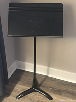 Black Manhasset professional music stand full view showing adjustable pole and three-legged base on wood floor.