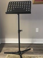 Front view of a black metal music stand with a perforated music holder panel, adjustable middle pole, and tripod base with legs extended on a wood floor against a plain wall.